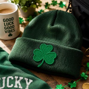 May include: A dark green knit beanie featuring a prominent embroidered shamrock patch on the front cuff. The hat is made of a ribbed material and is positioned near a white mug with the text GOOD LUCK GOOD VIBES and a small shamrock icon.