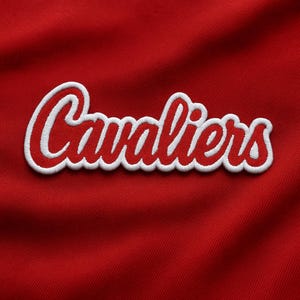 May include: Close-up of a red fabric with the word "Cavaliers" embroidered in white script with a red outline. The text is centered and slightly angled, with a textured appearance.