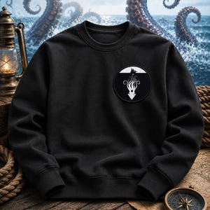 May include: A black crewneck sweatshirt featuring a circular patch on the left chest. The patch displays a white graphic of a sea creature with long tentacles extending downward from a small boat silhouette against a solid black background.
