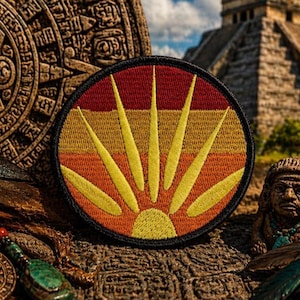 Puede incluir: Un parche bordado circular con un diseño de rayos de sol estilizado. El parche tiene un borde negro y un fondo de rayas horizontales en tonos rojo, naranja y amarillo, con rayos amarillos que irradian desde un sol central.