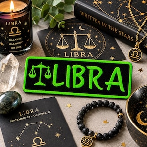 May include: A rectangular black patch with bright green embroidered text reading LIBRA and a scale symbol. The patch is surrounded by a Libra-themed candle, a black book titled WRITTEN IN THE STARS, a gold necklace, a black beaded bracelet, and cards.