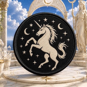 May include: A circular black embroidered patch featuring a cream-colored unicorn rearing on its hind legs, surrounded by a pattern of small stars and crescent moons. The design is stitched with a visible fabric texture and a thick black border.
