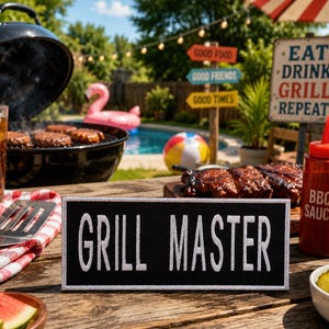 May include: A backyard barbecue scene with a grill, burgers, and ribs. A sign reads "GRILL MASTER." Condiments, watermelon, and a drink are on the wooden table. A pool and a sign that says "Eat Drink Grill Repeat" are in the background.
