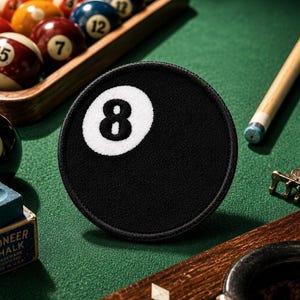 以下が含まれることがあります： 中央に黒い数字の8が描かれた白い円を持つ、円形の黒い刺繍ワッペン。ワッペンにはステッチの縁取りがあり、ビリヤードゲームの標準的な8番ボールの外観を模して直立した状態で配置されています。