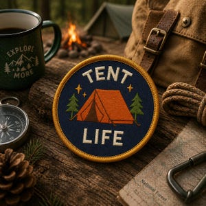 Puede incluir: Un parche bordado circular con fondo azul oscuro y un borde amarillo grueso. El parche presenta una tienda de campaña naranja, dos pinos verdes, dos estrellas amarillas y el texto blanco TENT LIFE centrado en la superficie de tela.