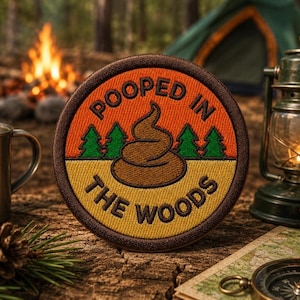 May include: A circular embroidered patch with a dark brown border. The top half is orange with green pine trees and the text POOPED IN. The bottom half is yellow with the text THE WOODS. A brown poop emoji icon is centred in the middle.