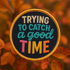 Puede incluir: Parche bordado redondo con el texto "TRYING TO CATCH a good TIME". El parche tiene un fondo negro con letras turquesas, rosas, naranjas y beige, y un borde dorado. El fondo es una imagen borrosa de follaje otoñal.