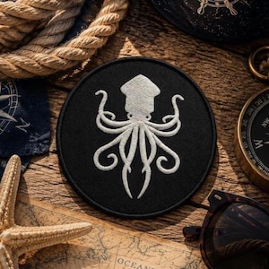 May include: A circular black embroidered patch featuring a white squid design with multiple curling tentacles. The patch has a stitched border and is centred, showcasing detailed thread work that forms the shape of the cephalopod creature.