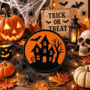 May include: A circular embroidered patch featuring a black silhouette of a haunted house, a bare tree, and a flying bat against a vibrant orange background. The patch has a black stitched border and is displayed among various Halloween decorations.