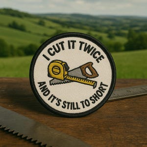 Könnte beinhalten: Runder Aufnäher mit schwarzem Rand und dem Text "I CUT IT TWICE AND IT'S STILL TO SHORT". Das Design zeigt ein gelbes Maßband und eine Säge mit braunem Griff. Der Hintergrund ist ein verschwommenes grünes Feld.