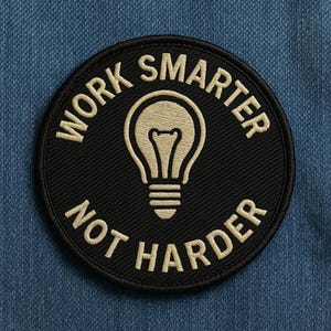 Może przedstawiać: Okrągła, czarna, haftowana naszywka z napisem "WORK SMARTER NOT HARDER" w kremowym kolorze. Kremowy symbol żarówki znajduje się na środku. Naszywka jest umieszczona na niebieskim tle z dżinsu.