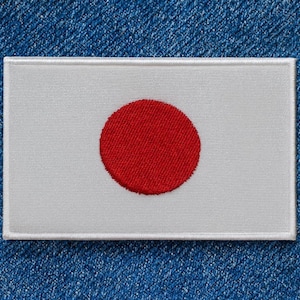 Puede incluir: Parche bordado con la bandera de Japón. El parche rectangular tiene un fondo blanco con un círculo rojo central, que representa el sol. El parche está bordeado con un borde blanco y está sobre un fondo de mezclilla azul.