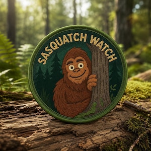 May include: A circular embroidered patch featuring a brown Sasquatch peeking from behind a tree trunk. The background is a dark green forest with silhouettes of pine trees. The text SASQUATCH WATCH is stitched in gold thread along the top edge.