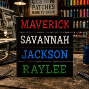 Puede incluir: Cuatro parches rectangulares negros bordados apilados verticalmente. Cada parche presenta un nombre en letras mayúsculas: MAVERICK en rojo, SAVANNAH en blanco, JACKSON en azul y RAYLEE en verde. El bordado es grueso y texturizado.