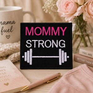 May include: A square black embroidered patch featuring the text MOMMY in pink and STRONG in white, with a white barbell graphic underneath. The patch has a clean, stitched border and is displayed as a standalone item for clear visibility.