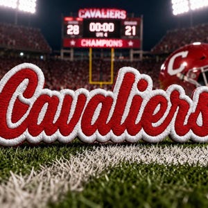 Puede incluir: Un parche bordado en rojo y blanco con la palabra Cavaliers en letra cursiva. El parche está colocado sobre un campo de césped verde con un fondo de estadio borroso que muestra un marcador con el texto Cavaliers y Champions.
