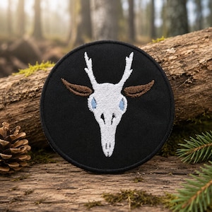 Könnte beinhalten: Ein kreisförmiger, schwarzer Stickaufnäher mit einem weißen Hirschschädel-Motiv und kleinem Geweih. Der Schädel ist an den Seiten mit braunen, ohrähnlichen Formen und hellblauen Details in den Augenhöhlen versehen, gestickt auf schwarzem Stoff.
