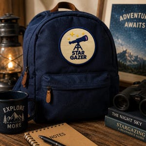 May include: A navy blue canvas backpack featuring a circular patch with a telescope graphic and the text STAR GAZER. The bag has a brown leather top handle and zipper pulls. The design is centered on the front pocket of the backpack.