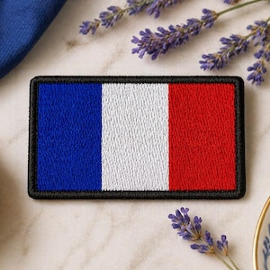May include: Embroidered patch featuring the French flag with blue, white, and red vertical stripes, framed by a black border. The patch is rectangular and sits on a marble surface, with lavender sprigs and a miniature Eiffel Tower nearby.