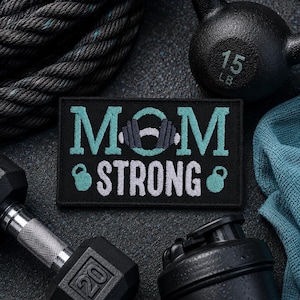 May include: A rectangular black embroidered patch featuring the text MOM STRONG in light blue and white thread. The letter O in MOM is replaced by a gray barbell graphic, and small light blue kettlebell icons flank the word STRONG.