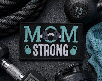 Può includere: Una toppa rettangolare nera ricamata con il testo MOM STRONG in filo azzurro e bianco. La lettera O è sostituita da un'icona di un bilanciere e piccole icone di kettlebell azzurre fiancheggiano la parola STRONG nella riga inferiore.