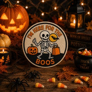Puede incluir: Un parche bordado circular que muestra un esqueleto blanco sosteniendo una calabaza naranja y una bolsa de dulces. El diseño incluye una luna amarilla con la silueta de un murciélago negro y el texto naranja I'M HERE FOR THE BOOS sobre fondo crema.