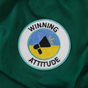 May include: A circular patch embroidered with the words "WINNING ATTITUDE". The design includes a black megaphone on a yellow and blue background, set against a green fabric.