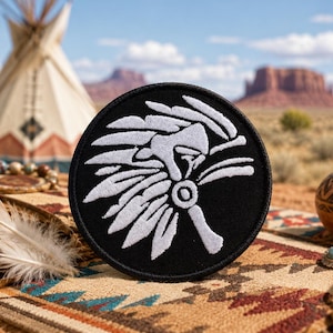 May include: A round, black patch with a white embroidered Native American headdress design. The patch is set against a backdrop of a desert landscape with a teepee, mountains, and decorative items, evoking a Southwestern theme.