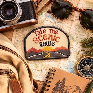 May include: An embroidered patch featuring a landscape with mountains, a winding road, and a yellow sun. The text reads Take The Scenic Route in red and black thread. The patch is surrounded by a camera, sunglasses, a map, a compass, and a notebook.