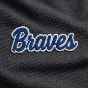 May include: Close-up of a dark gray fabric with the word "Braves" embroidered in blue with a white outline. The text is in a cursive font.