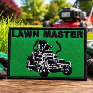 May include: A rectangular embroidered patch with a bright green background and a black border. The text LAWN MASTER is stitched in black at the top, and a black graphic of a lawn mower is centred below the text on the green fabric surface.