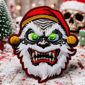 May include: An embroidered patch featuring a stylized, aggressive character with a white beard, sharp teeth, and bright green eyes. The figure wears a red hat with a gold trim and yellow pom-poms on the sides, outlined in a bold red border.