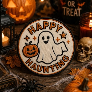 Puede incluir: Un parche bordado circular que presenta un fantasma blanco sosteniendo una pequeña calabaza naranja. El texto "HAPPY HAUNTING" está bordado en letras naranjas a lo largo de los bordes superior e inferior, acentuado por pequeñas estrellas naranjas sobre un fondo crema.