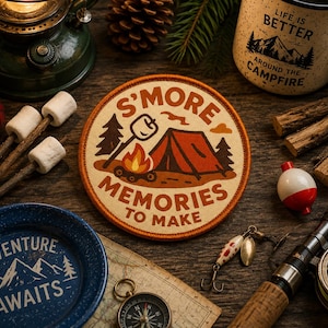 Puede incluir: Un parche con temática de camping con el texto "S'MORE MEMORIES TO MAKE" y un gráfico de una tienda de campaña, una fogata y árboles. La imagen también incluye una taza, aparejos de pesca, una brújula y un plato con la frase "ADVENTURE AWAITS".