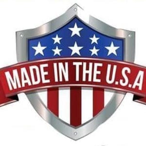 May include: A shield-shaped emblem featuring a blue field with white stars at the top and red and white vertical stripes at the bottom. A red ribbon banner overlays the center with the white text MADE IN THE U.S.A. in a bold, sans-serif font.