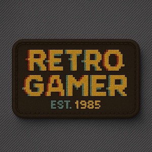 May include: A rectangular embroidered patch with a dark brown background and rounded corners. The text RETRO GAMER is stitched in a yellow pixelated font with red and blue shadow accents. Below, the text EST. 1985 is stitched in yellow.