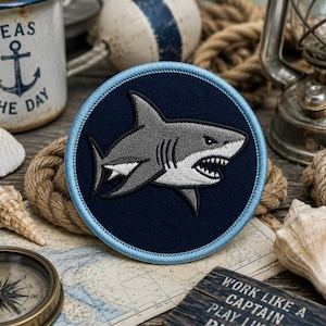 May include: A circular embroidered patch featuring a grey and white shark with an open mouth showing teeth, set against a dark navy blue background. The patch is finished with a light blue stitched border around the entire outer edge.