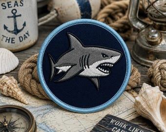Op de afbeelding: Een ronde geborduurde patch met een grijze en witte haai met een open mond en zichtbare tanden, geplaatst tegen een donkerblauwe achtergrond. De patch is afgewerkt met een lichtblauwe gestikte rand rondom de gehele buitenkant.