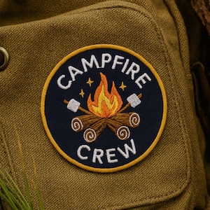 Könnte beinhalten: Ein kreisförmiger Aufnäher mit der Aufschrift "CAMPFIRE CREW", der ein Lagerfeuer-Design umgibt. Der Aufnäher befindet sich auf einem strukturierten olivgrünen Stoff. Das Lagerfeuer hat orange und gelbe Flammen, zwei Marshmallows und zwei Holzscheite.