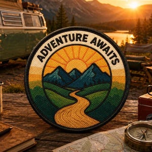 May include: A circular embroidered patch featuring a landscape design with a winding yellow path leading toward blue mountains under a yellow sun. The text ADVENTURE AWAITS is stitched in black along the top curve of the patch border.