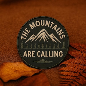 May include: A circular patch with the text "THE MOUNTAINS ARE CALLING" in white lettering. The patch features a mountain range graphic and a forest of trees. The patch is dark green with a black border. The background is brown.