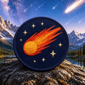 Puede incluir: Un parche bordado circular que presenta un cometa de colores naranja y amarillo vibrantes cruzando un fondo azul marino oscuro, rodeado por varias estrellas pequeñas de color dorado cosidas en un patrón circular sobre la tela.