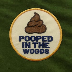 May include: Embroidered patch featuring a brown poop graphic on a white background, with a gold border. The navy blue text reads "POOPED IN THE WOODS". The patch is set against a green fabric.