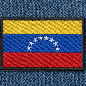Puede incluir: Parche bordado con la bandera de Venezuela. El parche rectangular tiene una parte superior amarilla, un centro azul con estrellas blancas y una parte inferior roja, todo delineado en negro. El parche está sobre un fondo de mezclilla azul.