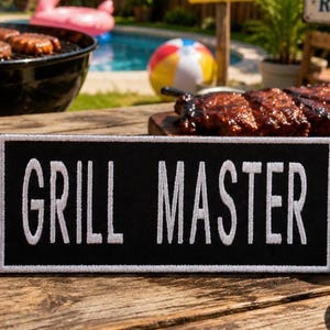 May include: A rectangular black patch with a white border and the white embroidered text "GRILL MASTER" in a clean, sans-serif font. The patch is positioned on a wooden surface, with a blurred outdoor barbecue setting in the background.