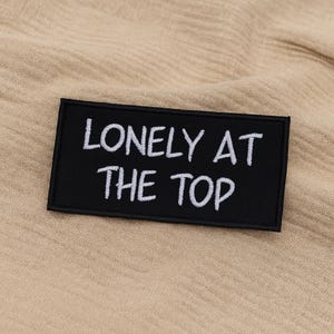 Pode incluir: Um patch retangular preto com o texto branco "LONELY AT THE TOP". O patch está em um fundo de tecido bege. O patch é provavelmente um patch para passar a ferro ou costurar em roupas ou acessórios.