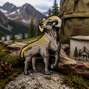 May include: Embroidered patch featuring a detailed depiction of a bighorn sheep. The patch showcases the animal in shades of gray, black, and yellow, with a black border. The background includes a mountain landscape, a backpack, and a camping mug.