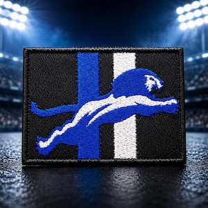 May include: A rectangular embroidered patch featuring a black background with a vertical blue stripe and a vertical white stripe. A stylised blue and white lion is depicted in a leaping motion across the centre of the patch, facing to the right.