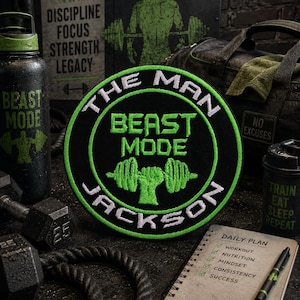 Peut inclure: Un écusson brodé circulaire avec un fond noir et une bordure vert vif. Le texte THE MAN BEAST MODE JACKSON est brodé en blanc et vert, accompagné d'un motif central représentant une main soulevant une barre de musculation en fil vert.
