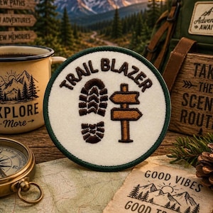 May include: A circular embroidered patch with a cream-coloured background and a dark green border. The patch features dark brown embroidery of a boot print, a wooden-style signpost, and the text "TRAIL BLAZER" arched across the top.